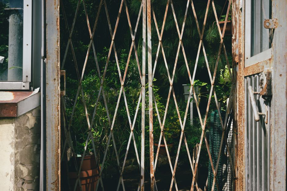 A close-up view of a rusted metal garden gate with a diamond lattice design, partially open to reveal a lush garden beyond. The garden features dense greenery with various plants and shrubs, some with